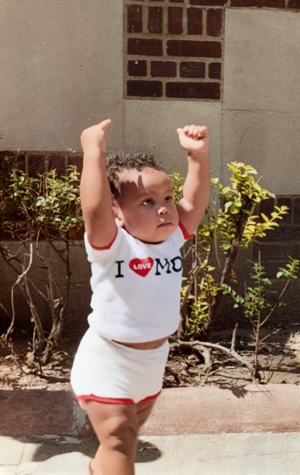 Kenny as a baby, arms raised proudly outside on a sunny day
